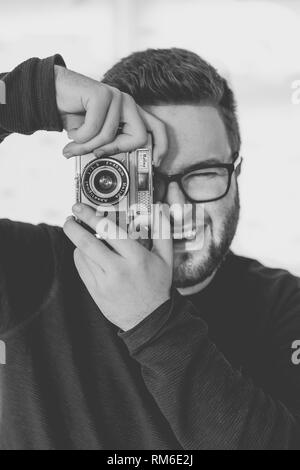 A  black and white portrait of a handsome, male, hipster photographer in glasses and taking photo with a vintage, 35mm camera whilst smiling Stock Photo