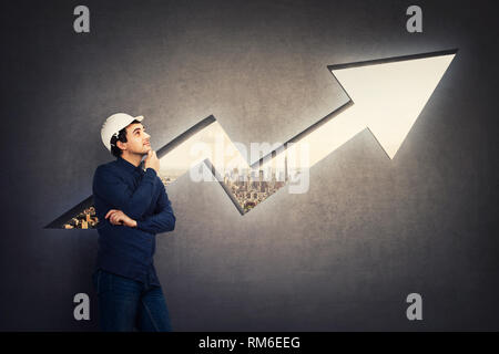 Wise engineer, wearing protective helmet, holding hand under chin thoughtful looking up in front of a rising graph arrow shaped as a hole in concrete  Stock Photo