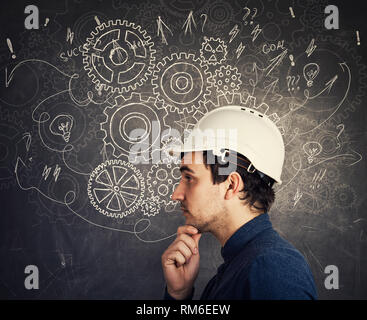 Hard thinking pensive engineer hand under chin looking thoughtful wearing protective helmet over blackboard with gear brain arrows and mess as thought Stock Photo