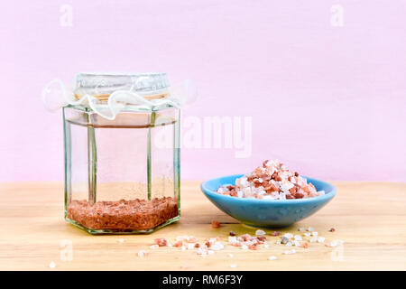 Himalayan salt sole, or sole water, in glass jar with a bowl of Himalayan salt, set on a wooden chopping board, against a pink background Stock Photo