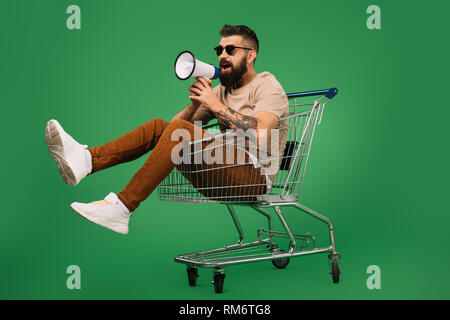 bearded man with megaphone sitting in shopping cart isolated on green Stock Photo