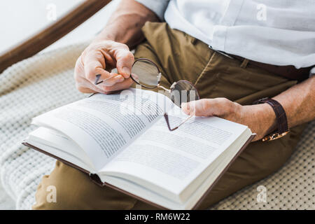 cropped view of senior man reading book and holding glasses at home Stock Photo