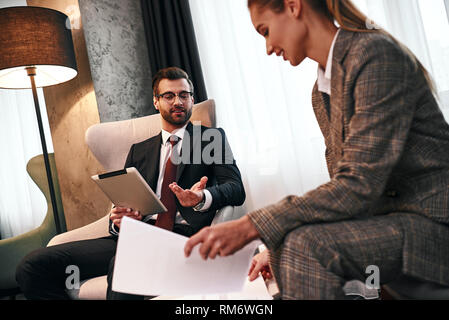 Contract details on tablet. Young smiling businessman talking with business woman and showing tablet and documents while having dinner in expensive hotel. Discuss contract details Stock Photo