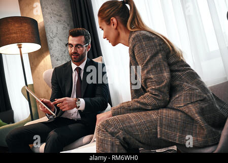 Look at this dashboard. Young smiling businessman talking with business woman and showing to her tablet while having dinner in expensive hotel. Discuss contract details Stock Photo