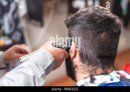 Handsome bearded man, getting haircut by barber, with electric trimmer at barbershop . Stock Photo