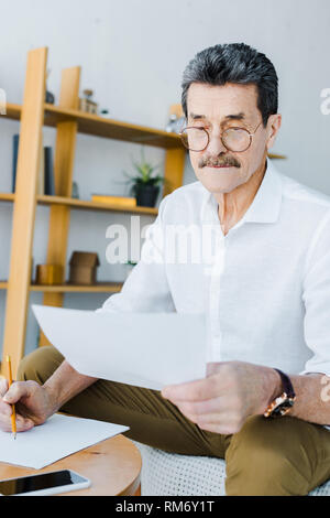 senior man in glasses holding pencil and looking at paper at home Stock Photo