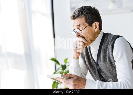 pensive retired man in glasses holding photo frame while sitting at home Stock Photo