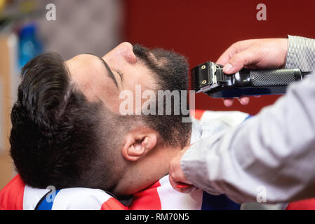 Barber shaving the beard of a handsome bearded man with an electric razor at the barber shop . Stock Photo