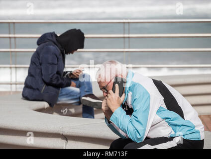 Elderly man and young man sitting on seat oveloooking the sea. Both using mobile phones Stock Photo