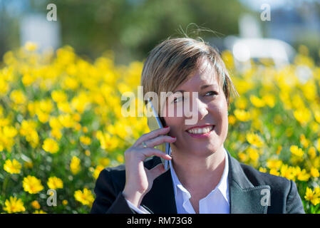 Front view of elegant smiling business woman sitting on a bench in a park while using a mobile phone enjoying the conversation outdoors Stock Photo