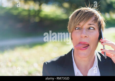 Front view of an elegant smiling business woman sitting on a bench in a park while using a mobile phone showing her tongue outdoors Stock Photo