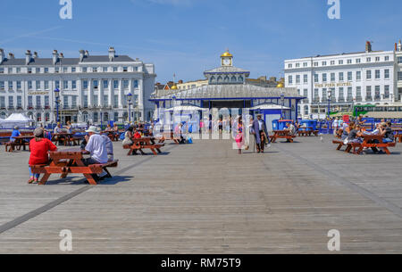 Eastbourne, Sussex, England, UK - August 1, 2018: People strolling and sitting on picnic benches near the entrance of the Pier in Eastbourne.  The Cla Stock Photo