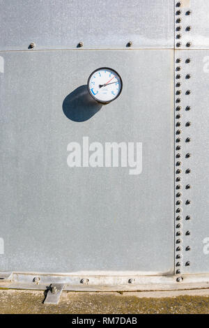 A round gauge fitted on the wall of a large galvanized steel water tank, with blue figures and two red and black needles on a white dial. Stock Photo