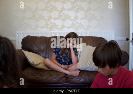 Bored looking child sits cross-legged on sofa, with older child(ren) in foreground. Rugby, England Stock Photo