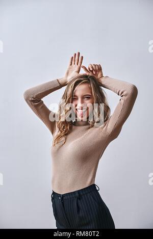 Portrait of positive flirty girl with brown hair dressed in beige turtleneck, looking happy, holding hands above her head, playfully. Looking at camera, sticking her tongue out, isolated over white background Stock Photo
