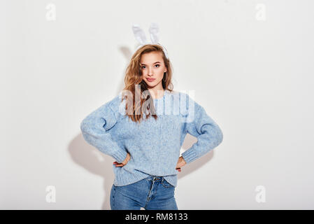 Portrait of playful thoughtful girl wearing blue sweater, jeans and bunny ears putting her hands on her hips, looking interestedly at camera, isolated over white background. Stock Photo