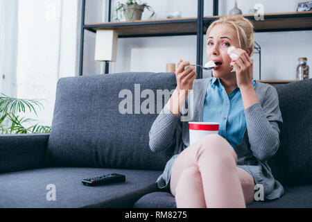 woman eating ice cream and crying while siitng on couch and watching tv at home alone Stock Photo