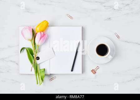 Office desk with coffee cup, paper clips, pencil, pink and yellow tulips with tag for name or message on open diary over white marble background. Flat Stock Photo