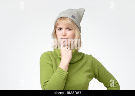 Blonde female wearing green sweater with puzzled expression, keeps finger on lips Stock Photo