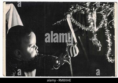 Retro photo shows toddler playing with christmas ball. Childhood and christmas tree. Black and white vintage photography. 1960s Stock Photo