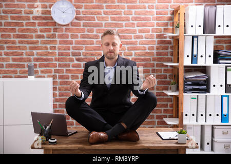 Young Businessman Sitting On Desk Meditating In Office Stock Photo