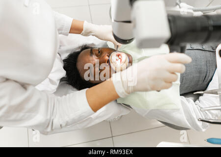 Young woman dentist treating root canals using microscope in the dental clinic. Man patient lying on dentist chair with open mouth. Medicine Stock Photo