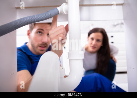 Young Male Plumber Showing Damage In Sink Pipe To Woman At Home Stock Photo