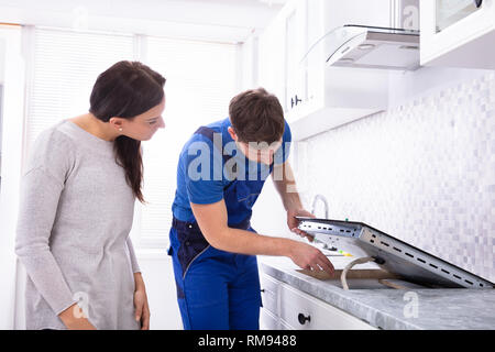 Woman Looking At Serviceman In Uniform Fixing Induction Stove In The Kitchen Stock Photo