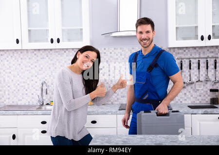 Smiling Young Woman Standing With Repairman Showing Thumbs Up Sign In Kitchen Stock Photo
