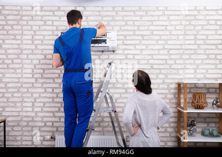 Smiling Woman Looking At Male Technician Repairing Air Conditioner With Digital Multimeter At Home Stock Photo