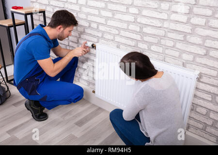 Beautiful Woman Looking At Young Male Plumber In Uniform Installing Radiator With Screwdriver Stock Photo