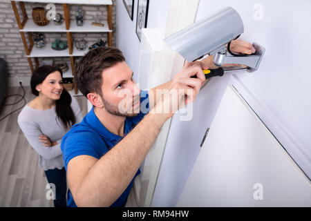 Smiling Woman Looking At Male Technician Adjusting CCTV Camera On Wall With Screwdriver Stock Photo