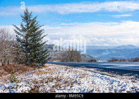 Winter landscape with wet melting road and hills with snow-covered trees, meadows and road shoulders and mountains hidden in the hanging clouds in the Stock Photo