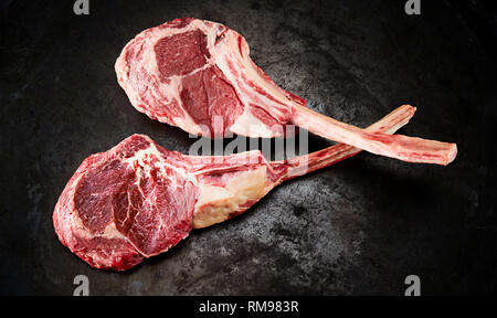 Two uncooked fatty ribeye or Delmonico steaks with the bone in viewed from above on an old textured blackened metal surface Stock Photo