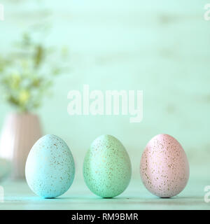Three colorful pastel Easter eggs decorated with golden splatter, flowers and spots in blue, green and yellow on mint green wood background with copy  Stock Photo