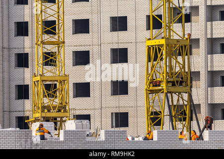 View of Construction Site With Unfinished Apartment Building, Cranes’ Bases and Construction Workers Dressed in Orange Outfit Stock Photo