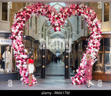 Burlington Arcade, Mayfair, London, UK. 14 February, 2019. Two young women stand at the entrance to Burlington Arcade in front of a giant pink heart on Valentines Day. Credit: Malcolm Park/Alamy Live News.