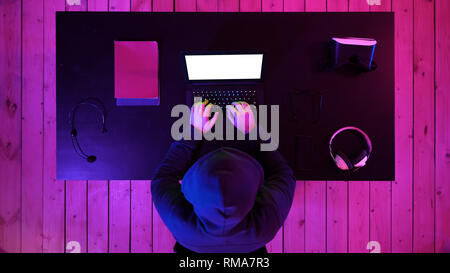 Hacker in hoodie typing code in program using laptop in dark studio. White Display. Stock Photo