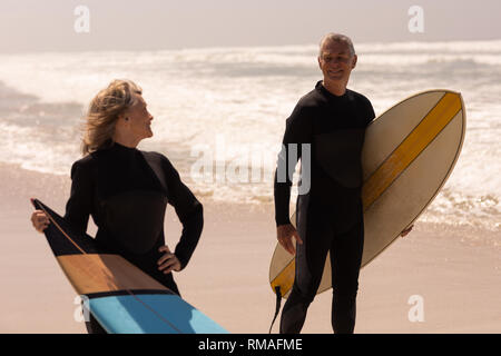 Senior couple with surfboard standing on beach and looking each other Stock Photo