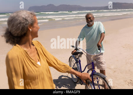 Senior couple standing with bicycle and looking each other on beach with mountains in the back Stock Photo