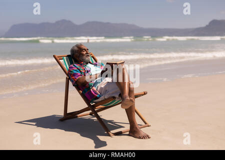 Senior man relaxing on sun lounger and reading a book on beach Stock Photo