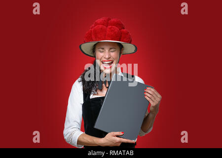 Candid portrait of a laughing attractive young Black Forest waitress holding a menu dressed in traditional clothes with a Bollenhut decorated with red Stock Photo