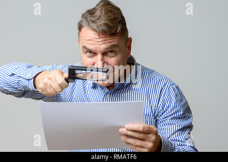 Middle aged business man using a magnifying glass to read a handheld document due to deteriorating eyesight, while looking to the camera Stock Photo