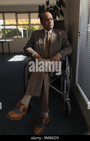 Front view of disabled senior man sitting on wheelchair and looking outside through window Stock Photo