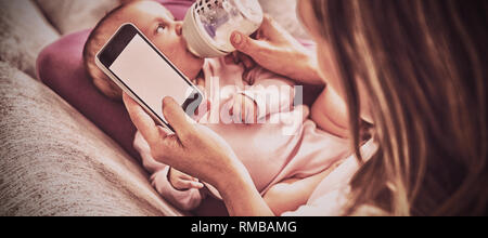 Mother using mobile phone while feeding her baby with milk bottle Stock Photo