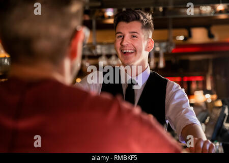Over the shoulder view of a young male bartender who is laughing and talking with a customer at the bar. Stock Photo