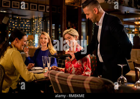 Mature waiter pouring a glass of prosecco for a table with three mature women sitting, laughing and smiling. Stock Photo