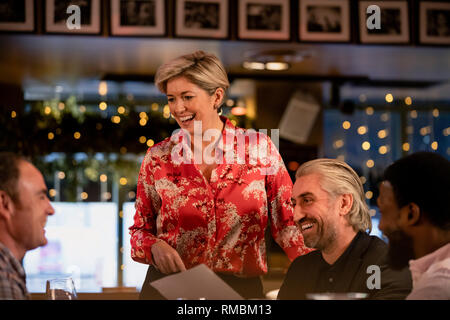 Smiling mature waitress taking an order of food from a small group of mid adult men in a restaurant. Stock Photo