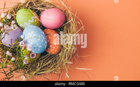 Easter eggs painted by children in a hay nest with flowers on a orange background. Top view Stock Photo