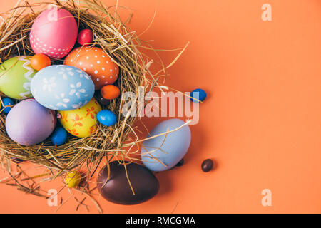 Easter eggs painted by children with chocolate and candies in a hay nest on a orange background. Top view Stock Photo